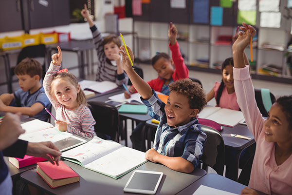 Young students sit in rows, raising their hands to answer the teacher