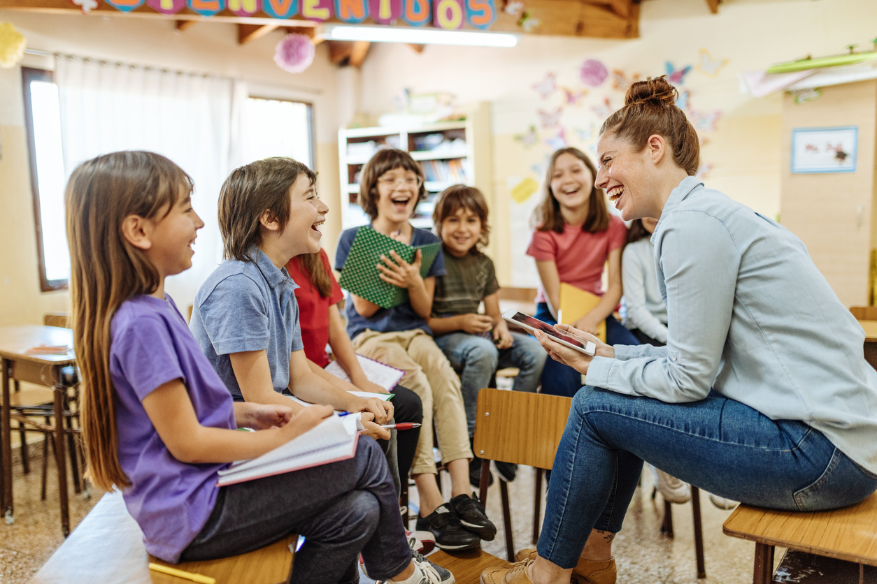 A teacher and seven of their students sitting on desks and laughing together.