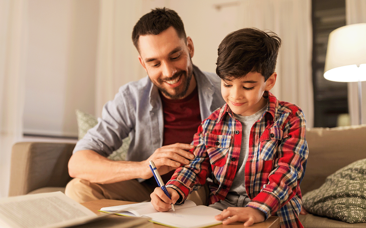  A father and a son sit together on a couch, focused on writing in notebooks